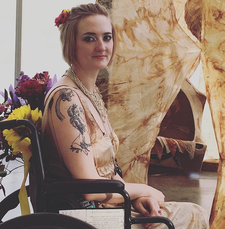 Robin Marquis looks directly at the camera with a slight smile on their face and bouquettes of flowers pour out of the back of their wheelchair. They are a light skinned white person with tattoos on their arm.